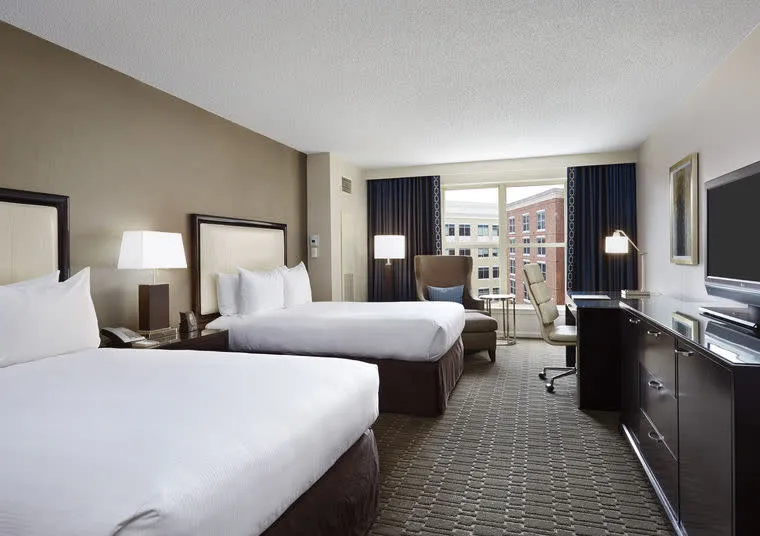 A hotel room with two large beds, white bedding, bedside lamps, a sofa chair by the window, a desk with a chair, a flat-screen TV, and a view of city buildings through large windows.