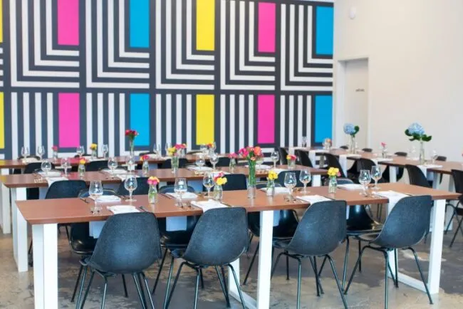 A modern dining area with wooden tables, black chairs, and place settings, featuring colorful flowers in vases. The background wall has a bold geometric pattern in black, white, blue, pink, and yellow.