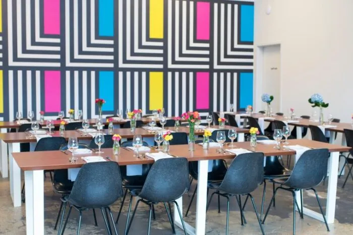 A modern dining area with wooden tables, black chairs, and place settings, featuring colorful flowers in vases. The background wall has a bold geometric pattern in black, white, blue, pink, and yellow.
