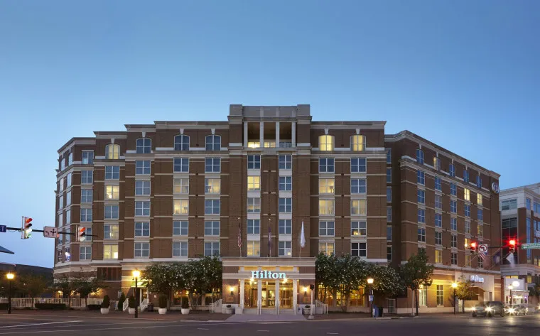 A multi-story Hilton hotel building with large windows, warm lighting inside, and a symmetrical brick facade, situated on a city street at dusk. Traffic lights and trees are visible in front of the entrance.