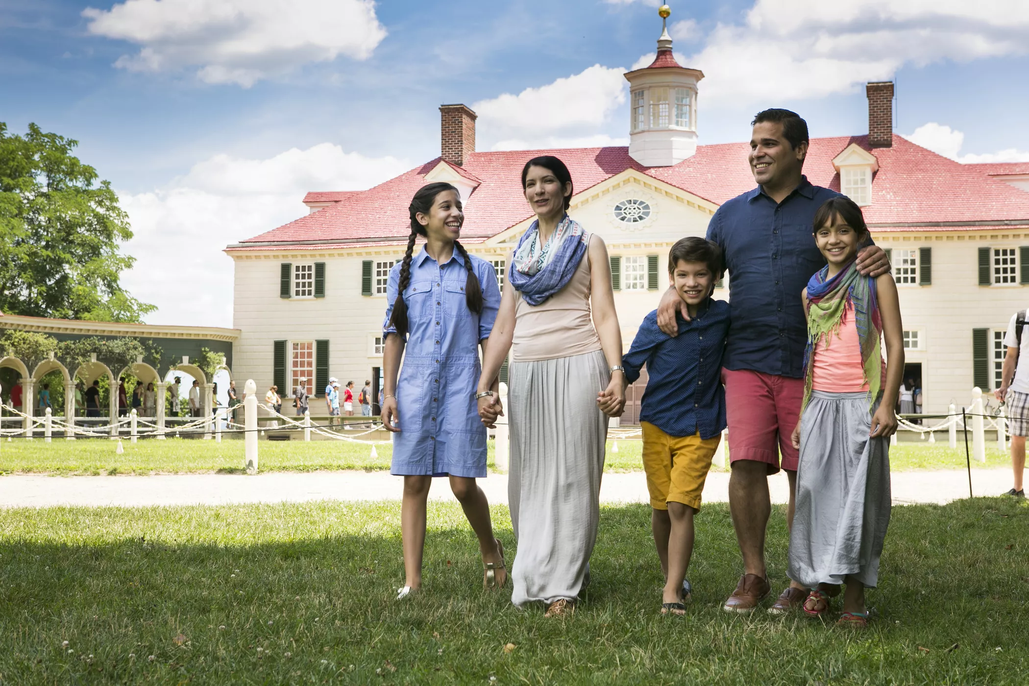 A smiling family of five walks on grass in front of a large historic building with a red roof and cupola under a blue sky with clouds. The parents hold hands with their three children, enjoying a sunny day.