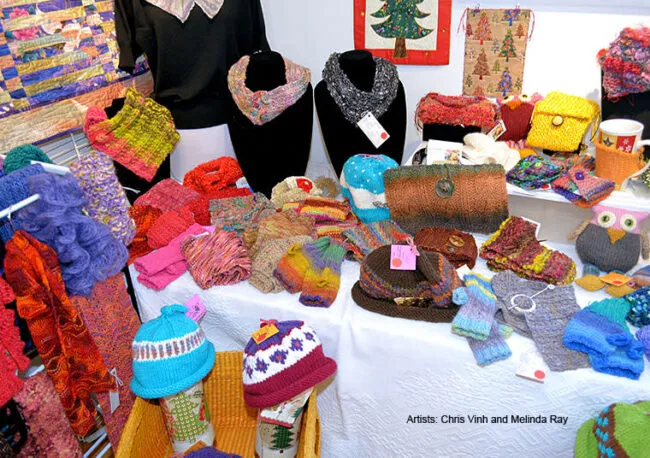 A colorful display of handmade knitted accessories, including hats, scarves, mittens, purses, and small pouches, arranged on a white tablecloth at a craft fair. Text in the image credits artists Chris Vinh and Melinda Ray.