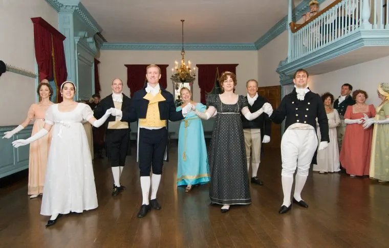 A group of people in Regency-era costumes hold hands in pairs, dancing in a historic ballroom with wooden floors, red curtains, and chandeliers. Some observers watch from a balcony above.
