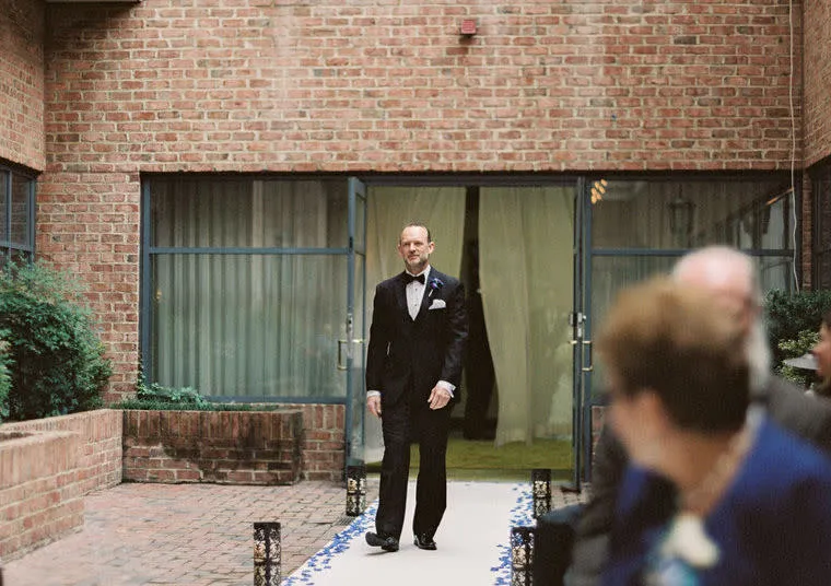 A man in a dark suit walks down an aisle covered with white fabric and flower petals in an outdoor brick courtyard, while seated guests watch in the foreground.