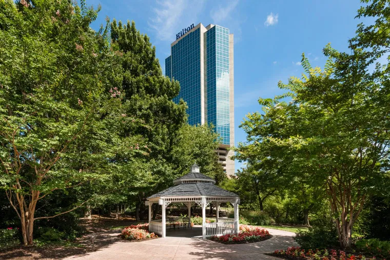 A white gazebo surrounded by trees and colorful flowers, with a tall glass Hilton hotel building in the background under a blue sky.
