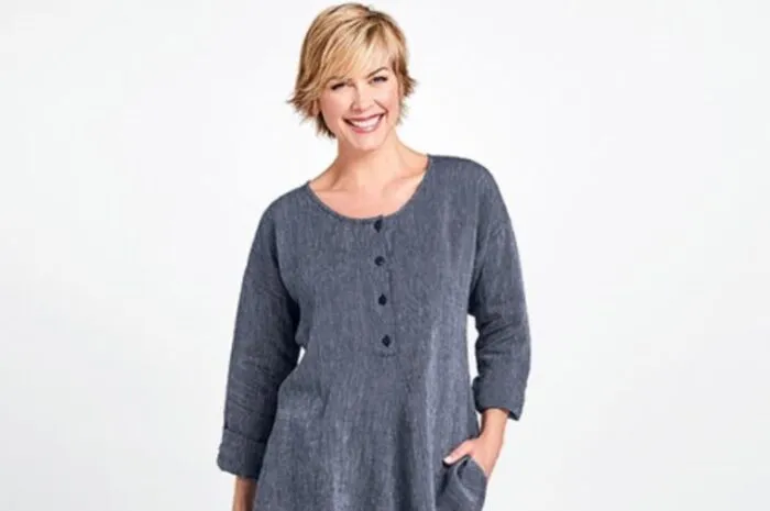 A smiling woman with short blonde hair wearing a loose-fitting, long-sleeve, buttoned gray dress stands with one hand in her pocket against a plain light background.