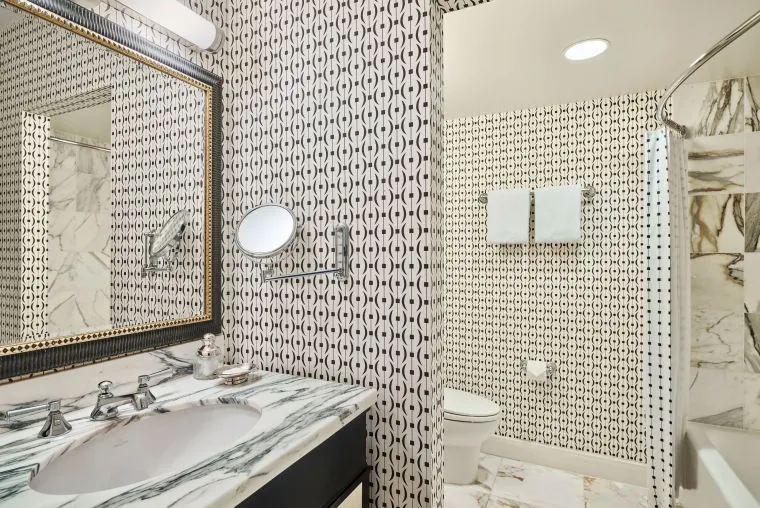 A modern bathroom with black and white geometric-patterned wallpaper, a marble countertop and sink, wall-mounted mirror, toilet, and glass shower. Marble tiles appear on the floor and in the shower area.