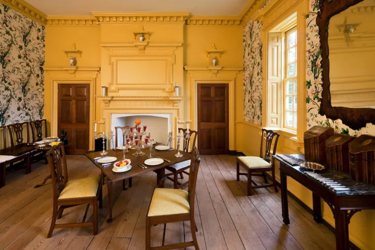 A historic dining room with yellow walls, a decorative fireplace, wooden floors, and windows. A table set for four with plates and glasses, surrounded by wooden chairs, sits in the center. A mirror and sideboard are on the right.