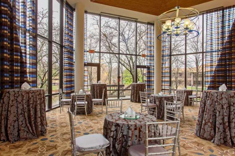 A bright event space with tall glass windows, round tables covered in patterned cloths, folding silver chairs, and views of trees outside. The room has patterned carpet, striped curtains, and a modern chandelier overhead.