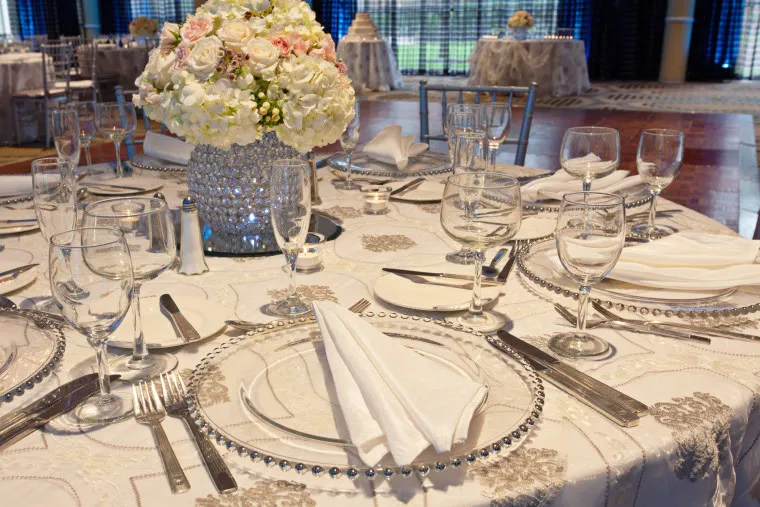 A round banquet table elegantly set with glass plates, silverware, folded white napkins, and clear glassware. The centerpiece is a sparkling vase with white and pale pink flowers. The room appears ready for a formal event.