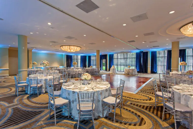 Elegant banquet hall with round tables covered in white cloths, silver chairs, floral centerpieces, and a wooden dance floor. Large windows line the back, and blue accent lighting brightens the modern space.