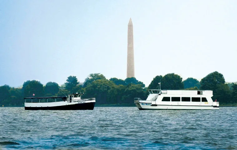 Two boats float on a river with green trees in the background. The Washington Monument rises in the distance under a hazy blue sky.
