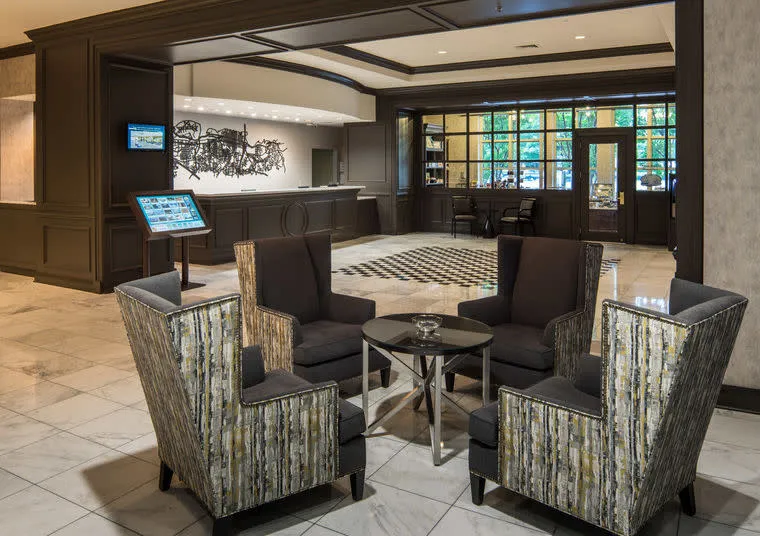 A hotel lobby with four patterned armchairs around a small table, marble flooring, a reception desk in the background, and large windows letting in natural light.