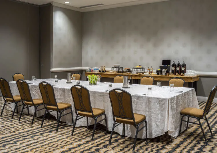 A conference room with a U-shaped table setup covered in white tablecloths, surrounded by tan chairs. On the side, a refreshment table holds coffee, drinks, and snacks. The room has patterned carpet and neutral walls.