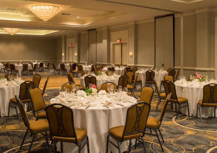 A banquet hall with round tables covered in white tablecloths, set with glassware and silverware. Each table has several brown chairs and a small floral centerpiece. The room is well-lit with chandeliers.