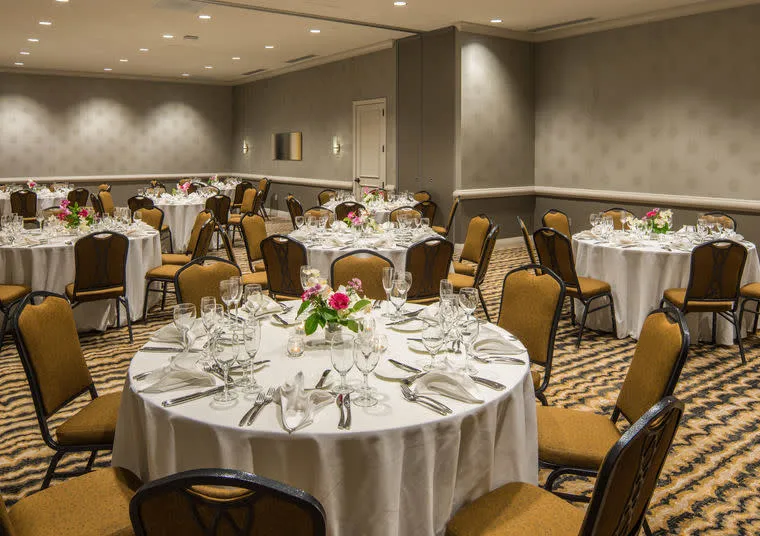 A banquet hall set for an event with round tables covered in white cloths, each decorated with pink and white floral centerpieces, neatly arranged glassware, and cutlery. Brown cushioned chairs surround each table.