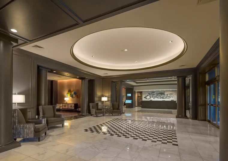 Spacious hotel lobby with marble floors, black and white checkered tile design, modern seating areas, columns, soft lighting, and a reception desk in the background.