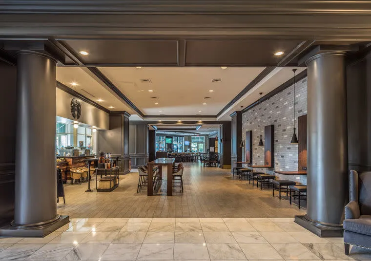 Spacious, modern restaurant interior with tall columns, marble floors, wooden tables and chairs, pendant lighting, and large windows at the far end letting in natural light.