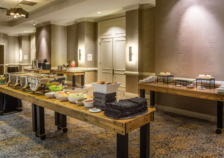 A buffet setup in a well-lit room with wooden tables displaying salads, chafing dishes, pastries, and desserts, arranged neatly against beige walls and patterned carpet.