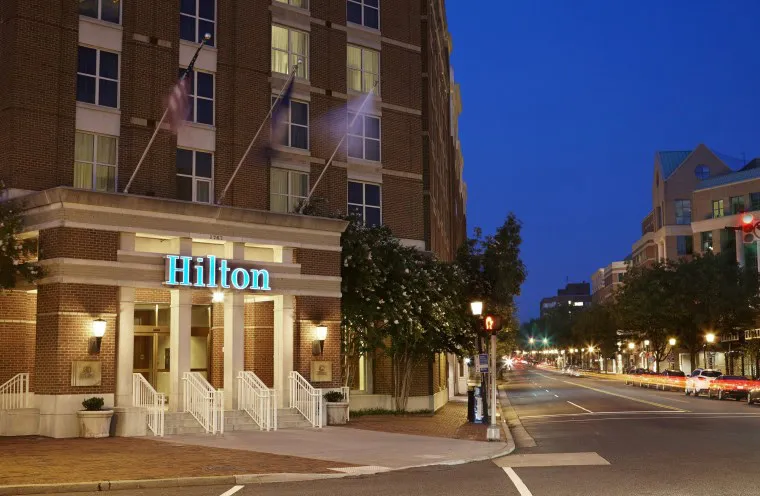 A Hilton hotel building with illuminated signage at night, located on a quiet, tree-lined street with parked cars and lit street lamps. The sky is dark blue, and the hotel entrance is well-lit.