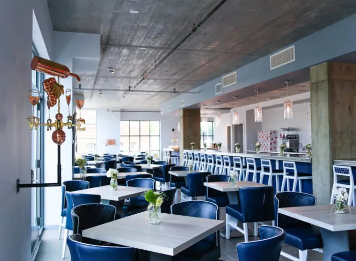 Modern restaurant interior with blue chairs, white tables, and small floral centerpieces. A bar with high white stools runs along the right side, and a wine-themed wall decor is mounted on the left. Large windows let in natural light.