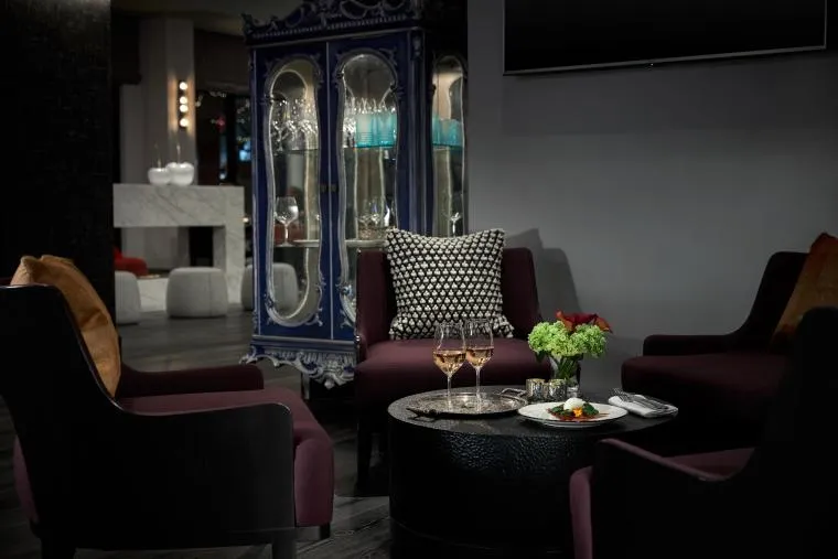 Four dark armchairs surround a round table with two glasses of wine, a plate of food, and a small flower arrangement. In the background is an ornate blue display cabinet and modern decor. The setting appears elegant and cozy.