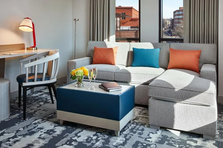Modern living room with a gray sectional sofa, orange and blue accent pillows, a blue ottoman as a coffee table with flowers and drinks, a desk with a chair, and large windows letting in natural light.