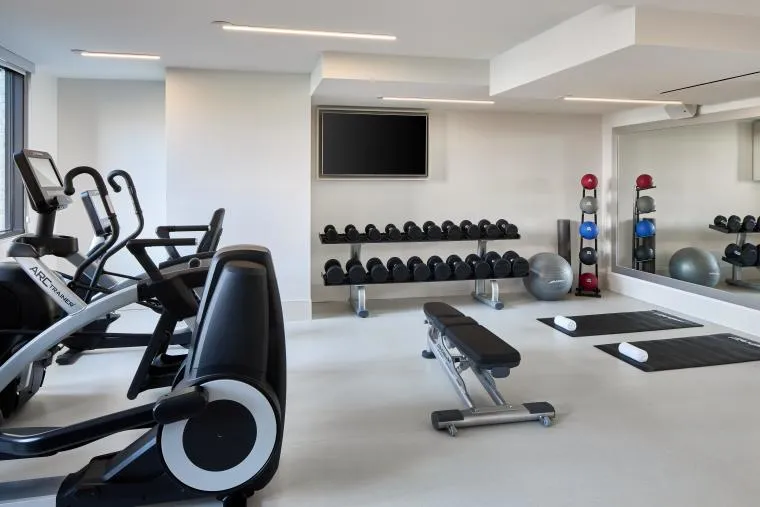 A modern gym with exercise bikes, a weight bench, dumbbells on a rack, stability balls, yoga mats, and a large wall mirror. A flat-screen TV is mounted on the wall above the weights.