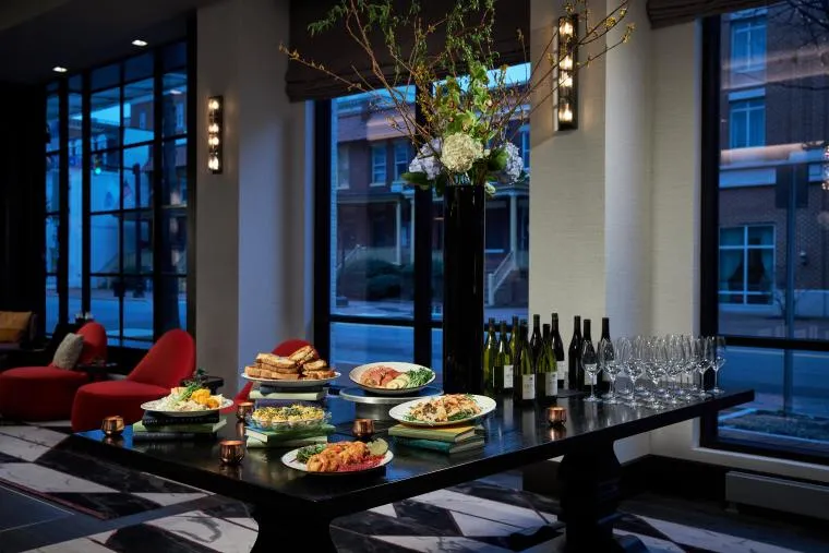 A black table set with plates of assorted food, bottles of wine, and empty wine glasses stands in a modern room with large windows and red chairs. A tall floral arrangement decorates the table’s center.