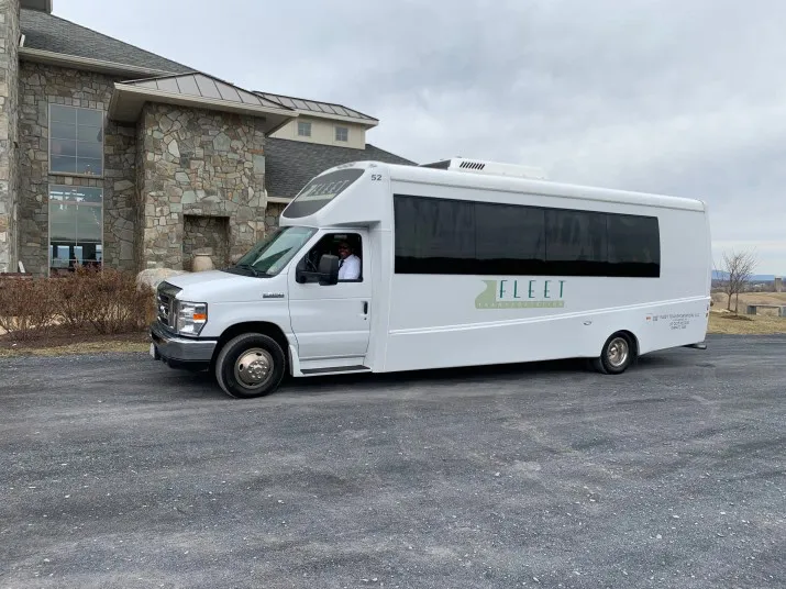 A white shuttle bus labeled Fleet is parked on a paved surface near a stone building under an overcast sky. A person is visible in the drivers seat.