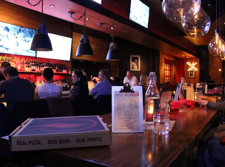 A lively bar and restaurant with people seated at the counter. Menus and a pizza box are on the wooden bar, with hanging lights above and bottles lining the shelves in the background. TV screens display images on the wall.