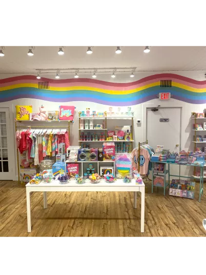 A colorful boutique with rainbow-striped walls, children’s clothing, toys, and gifts neatly arranged on shelves and a central table, all set on a wood floor under bright ceiling lights.