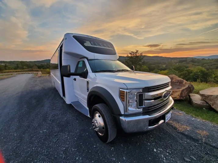 A white shuttle bus is parked on a gravel road at sunset, with green hills and trees in the background under a partly cloudy sky.