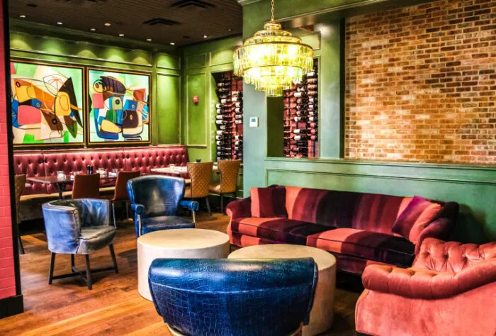 A vibrant restaurant lounge with colorful abstract art, plush velvet seating in red and blue tones, a green wall, exposed brick, a chandelier, and a wine rack display.
