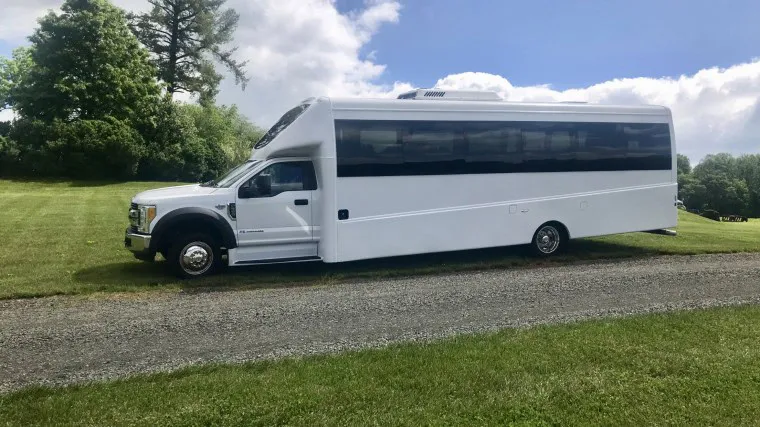 A large white shuttle bus is parked on grass next to a gravel road, with green trees and a partly cloudy sky in the background.