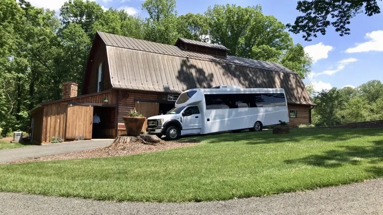 A large white shuttle bus is parked on a driveway in front of a rustic wooden barn surrounded by green trees and grass on a sunny day.