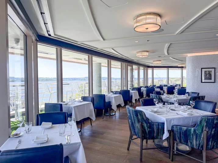 A bright, modern restaurant with blue chairs and white tablecloths. Large windows offer a scenic view of water and greenery outside. Tables are elegantly set for dining, and natural light fills the space.