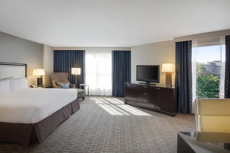 Spacious hotel room with a king bed, armchair, TV on a dark wood dresser, large windows with blue curtains, a beige lounge chair, and carpeted floor. Sunlight streams in through the windows.