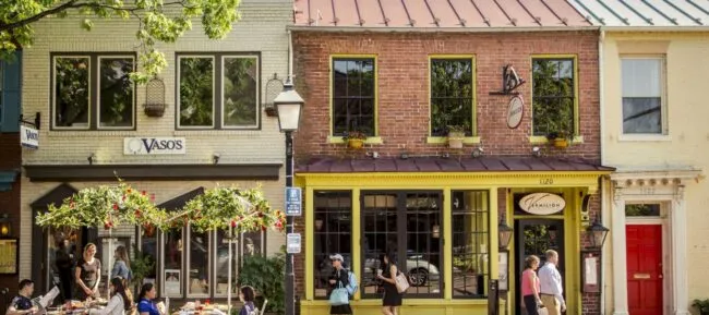 People dine outdoors at a café on a sunny day in front of charming brick and painted storefronts on a quaint street, with trees and a vintage-style lamppost nearby.