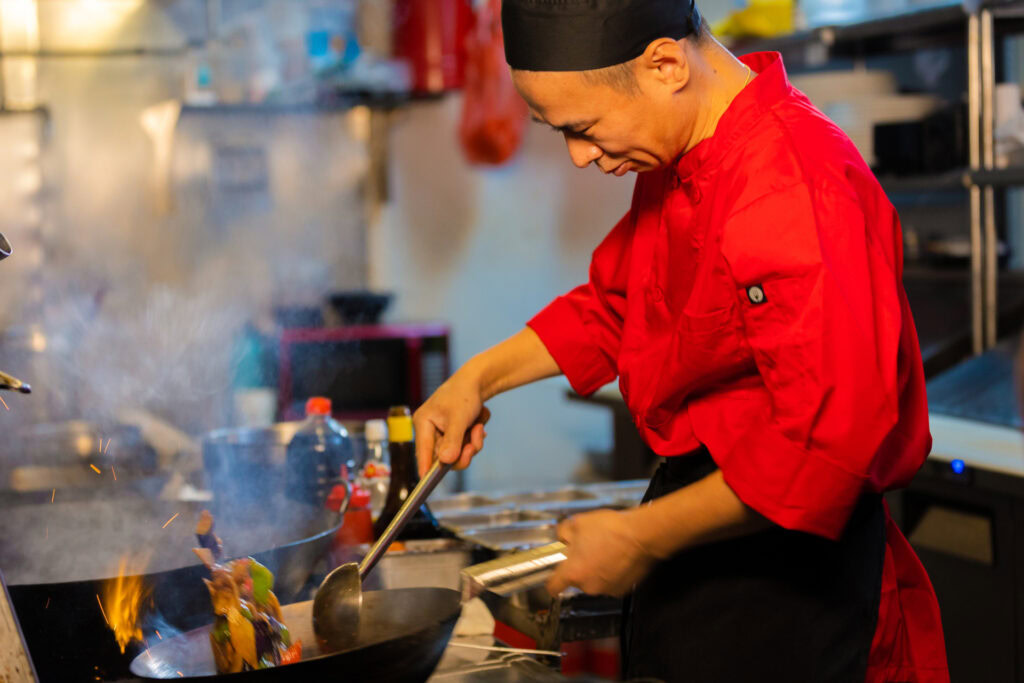 A chef in a red uniform stir-fries vegetables in a wok over a high flame in a commercial kitchen, focused on cooking as steam rises from the pan.