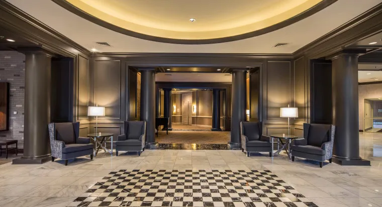 Elegant hotel lobby with marble floors, black and white checkerboard pattern, dark columns, high ceilings, two pairs of armchairs with side tables and lamps, and a softly lit entryway in the background.
