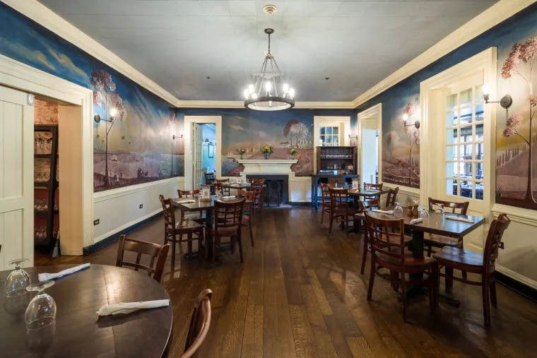 A cozy, historic dining room with wooden floors, several tables and chairs, a fireplace, mural-painted walls, and a chandelier hanging from the ceiling. Daylight comes through large windows on the right side.