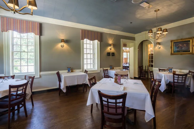 A bright, elegant dining room with several tables covered in white tablecloths, wooden chairs, large windows letting in natural light, and wall sconces providing additional lighting.