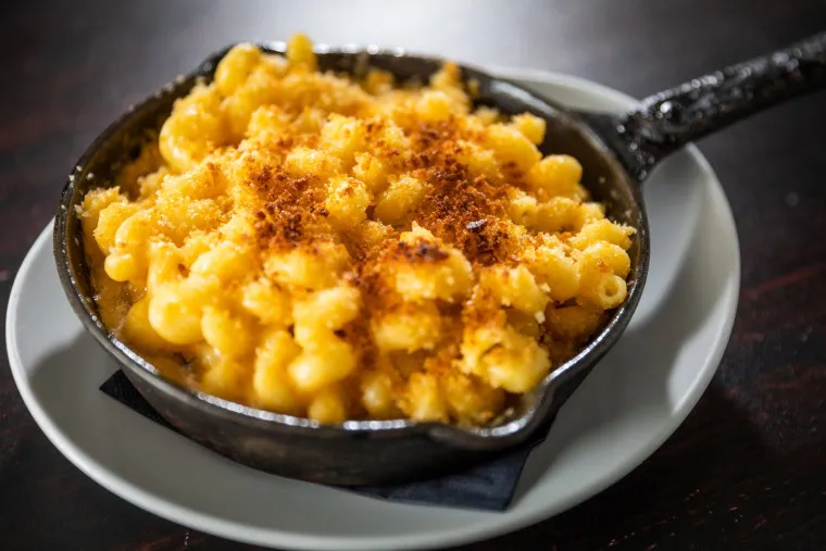 A small cast iron skillet filled with baked macaroni and cheese, topped with golden brown breadcrumbs, sits on a white plate against a dark background.