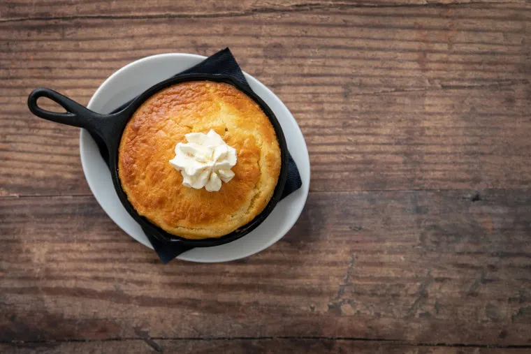 A small skillet with a golden-brown baked dessert topped with a dollop of whipped cream, placed on a white plate and set on a rustic wooden table.