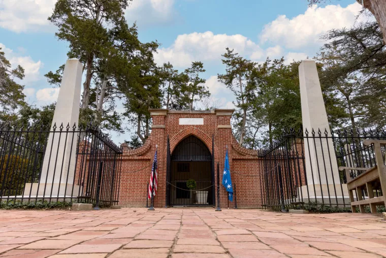 A brick mausoleum with arched entrance, flanked by two tall stone pillars and black iron fencing; American and blue flags stand outside, with trees and a partly cloudy sky in the background.