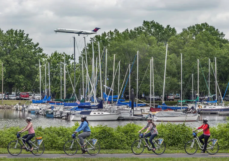 Four people ride bikes on a path beside a marina filled with sailboats. Behind them, a large airplane is flying low overhead, approaching to land. Trees and a cloudy sky are in the background.