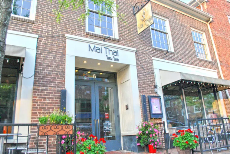 Front view of Mai Thai Restaurant, with a brick facade, blue double doors, large windows, outdoor seating, potted plants, and a hanging sign above the entrance.