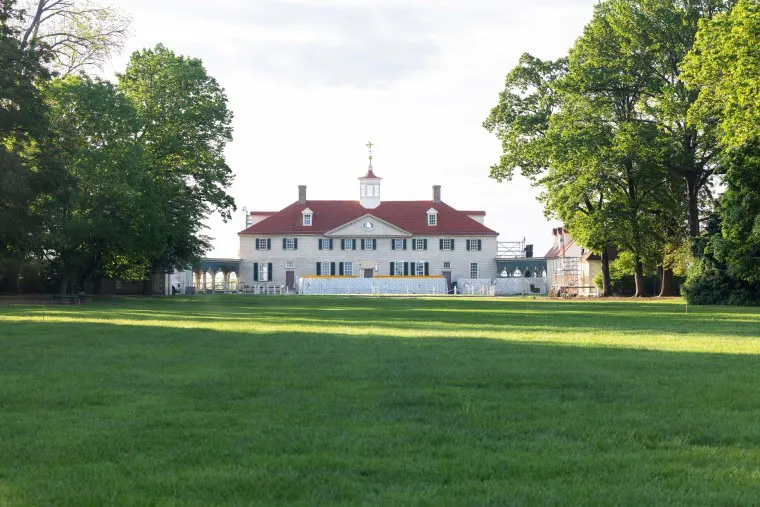 A large historic mansion with a red roof and white walls stands at the end of a wide, green lawn, surrounded by tall leafy trees under a bright sky.