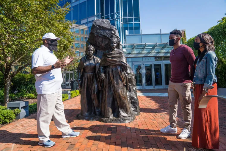 A man speaks to two people in front of a large bronze statue of two women outside on a brick walkway, with trees and a modern glass building in the background. All three people are wearing face masks.